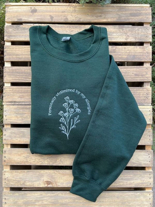 A dark green sweatshirt that says "personally victimized by my allergies" in a half circle over a branch of flowers. Line art design. White text. Shirt is on wooden slats with the left sleeve displayed flat.