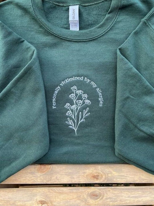 A dark green sweatshirt that says "personally victimized by my allergies" in a half circle over a branch of flowers. Line art design. White text. Shirt is laid out flat.