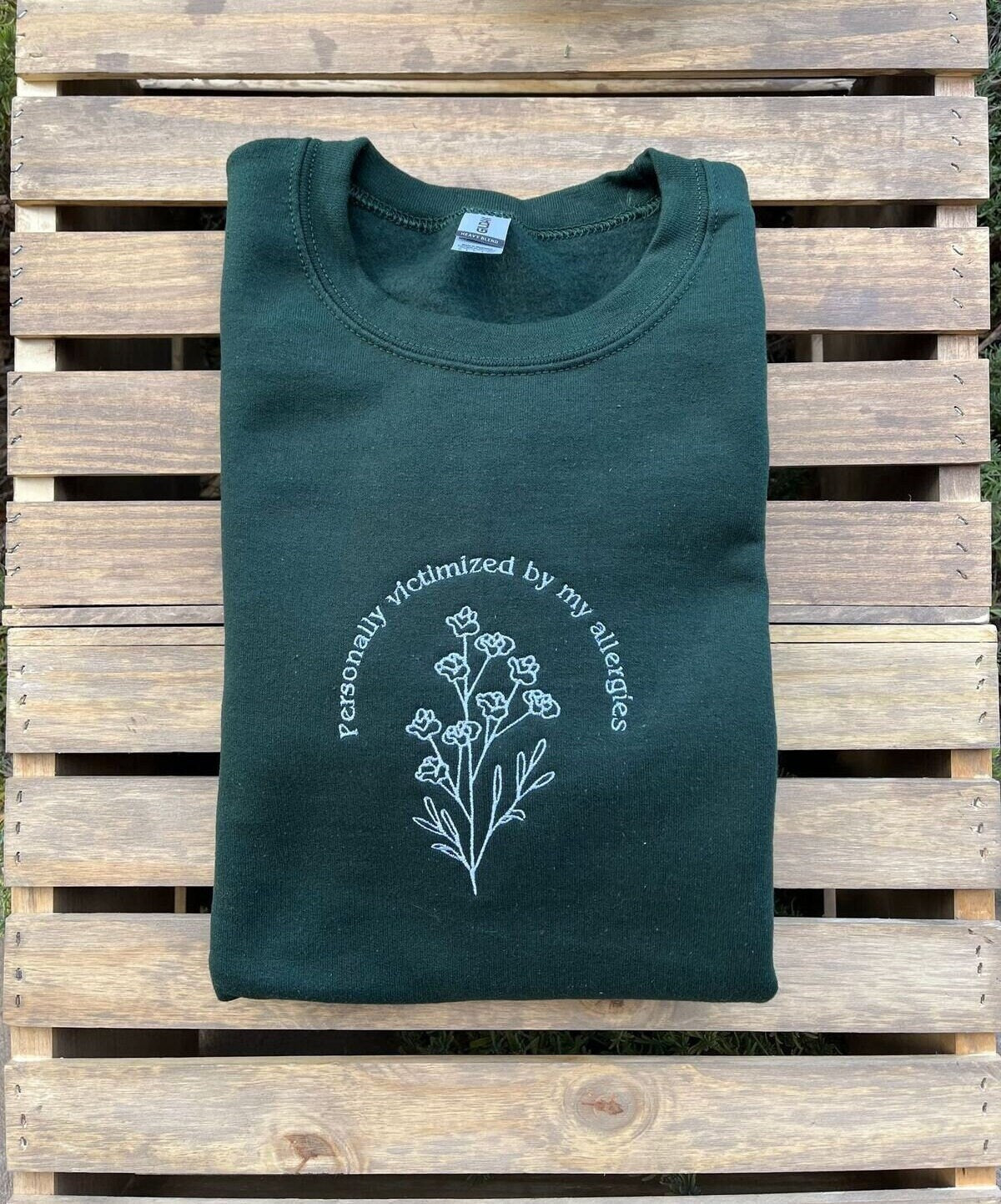 A dark green sweatshirt that says "personally victimized by my allergies" in a half circle over a branch of flowers. Line art design. White text. Shirt is folded up on some wooden slats.