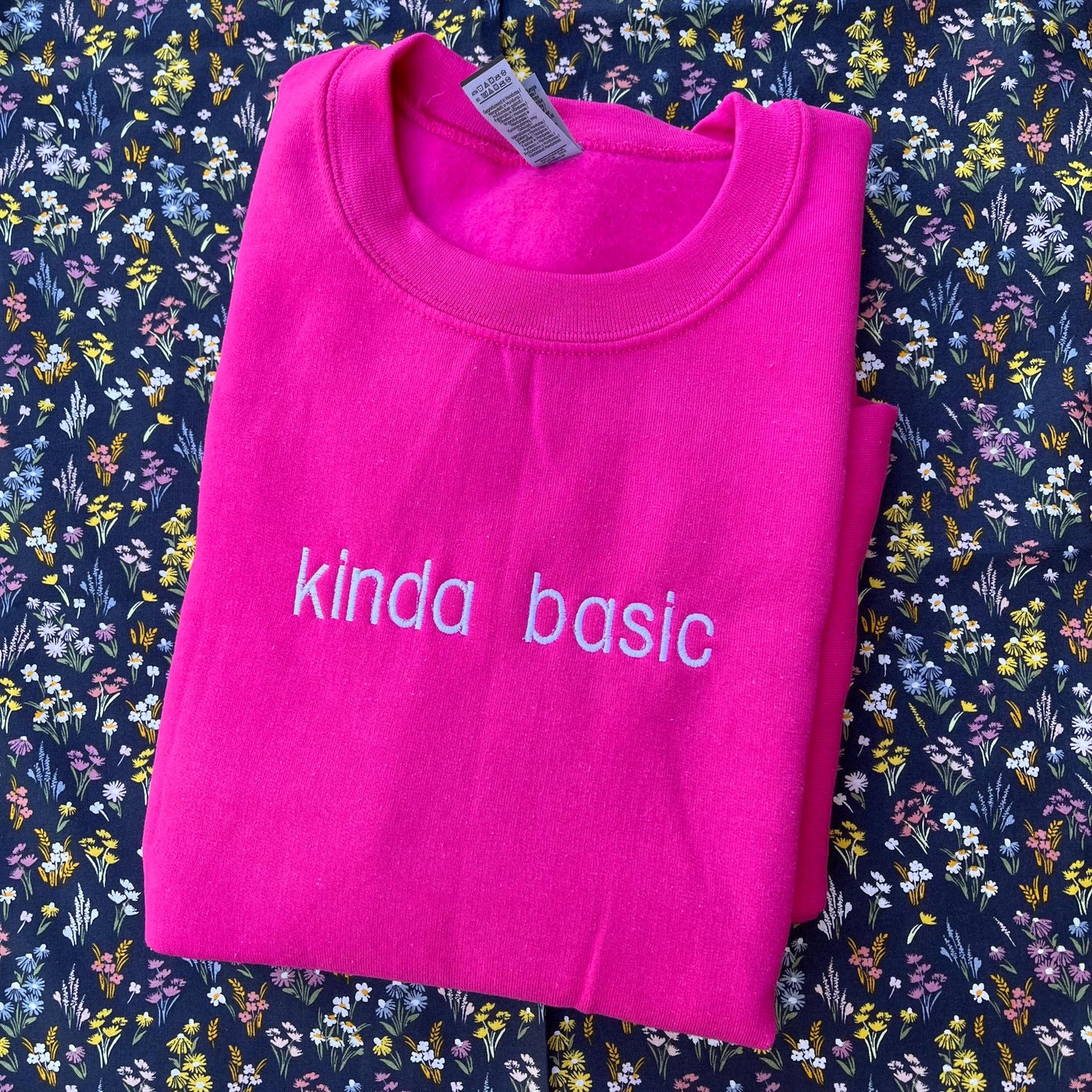 The image shows a folded bright pink sweatshirt with the words "kinda basic" embroidered in white on the front. The sweatshirt is placed on a floral-patterned fabric background with small, colorful flowers.