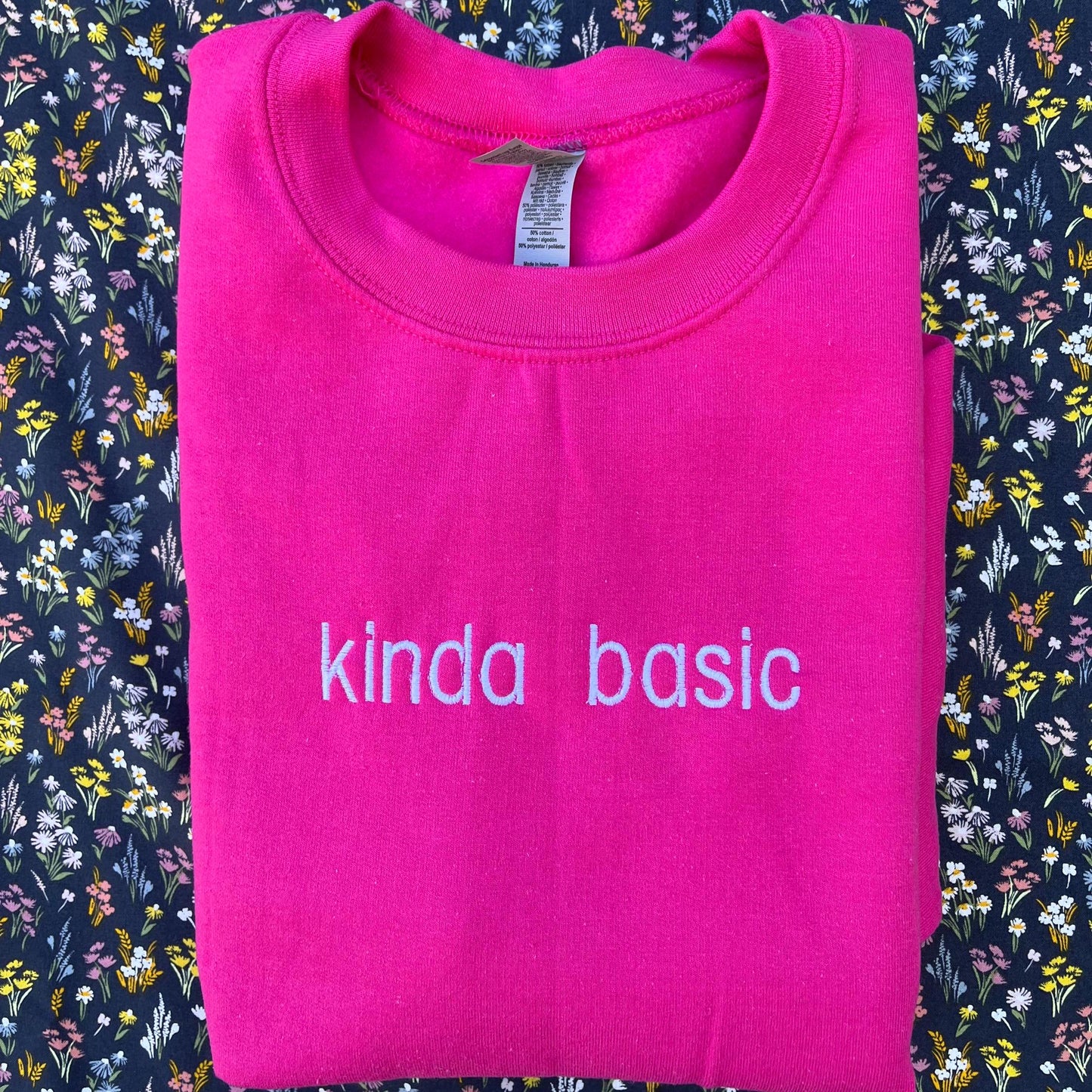 The image shows a folded bright pink sweatshirt with the words "kinda basic" embroidered in white on the front. The sweatshirt is placed on a floral-patterned fabric background with small, colorful flowers.