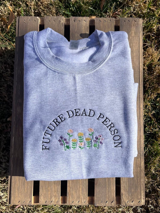 A gray embroidered sweatshirt with the words &quot;Future Dead Person&quot; in a semicircle. Multicolored flowers are under the lettering. The sweatshirt is displayed on wooden slats.