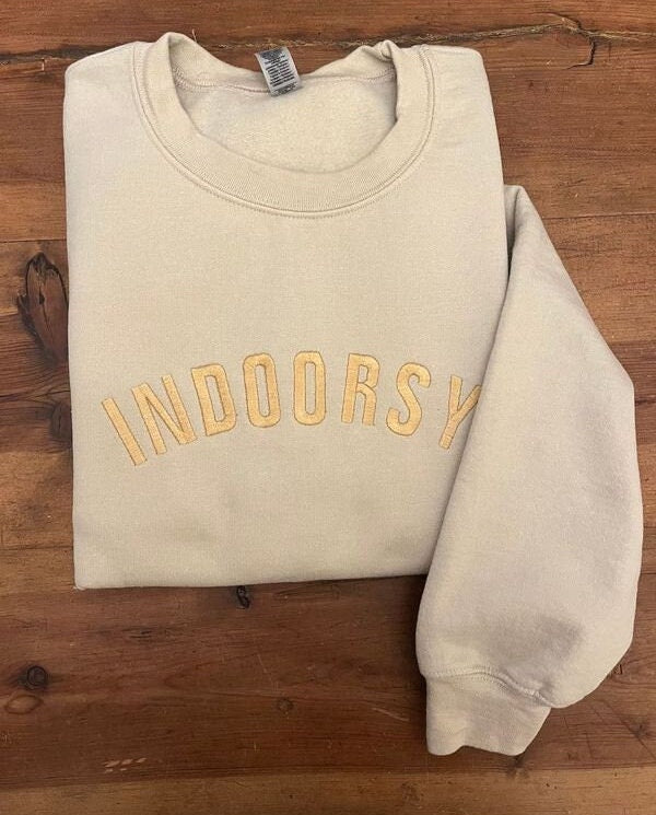 Sand sweatshirts with indoorsy in all caps across the front. It has gold lettering. Left arm is displayed.