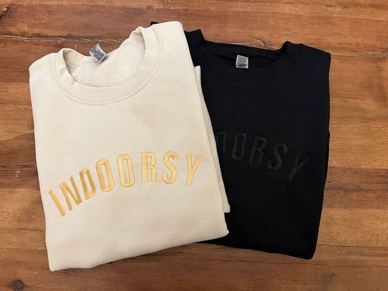 Black and sand sweatshirts with indoorsy in all caps across the front. Sand sweatshirt has gold lettering and black sweatshirt has black lettering.