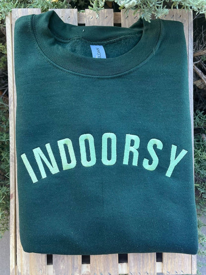 Forest green sweatshirt with indoorsy in all caps across the front in light green. Shirt is folded on wooden slats.