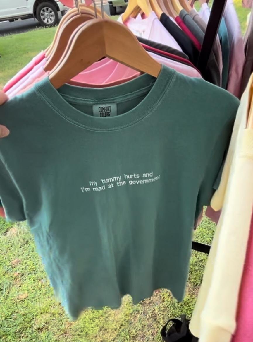 The image shows an emerald green shirt with the text “my tummy hurts and I’m mad at the government” printed in small white letters on the front. The shirt is hanging on a wooden hanger on a clothing rack with other apparel outside.