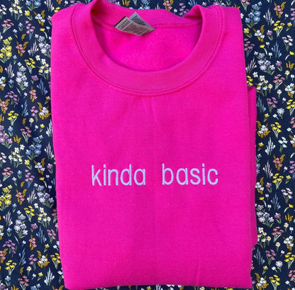 The image shows a folded bright pink sweatshirt with the words &quot;kinda basic&quot; embroidered in white on the front. The sweatshirt is placed on a floral-patterned fabric background with small, colorful flowers.