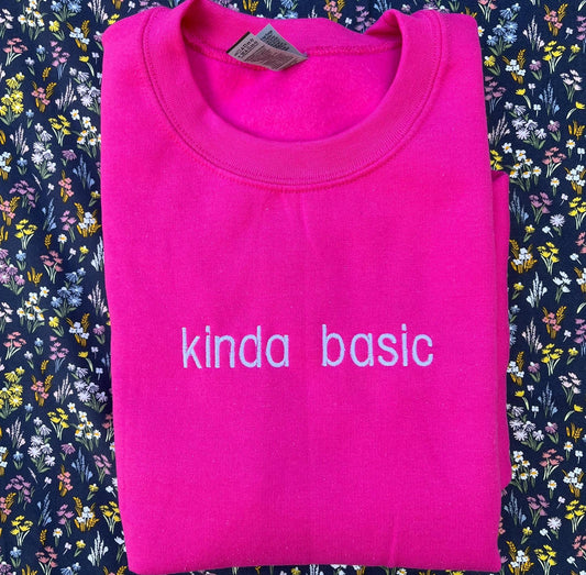 The image shows a folded bright pink sweatshirt with the words &quot;kinda basic&quot; embroidered in white on the front. The sweatshirt is placed on a floral-patterned fabric background with small, colorful flowers.