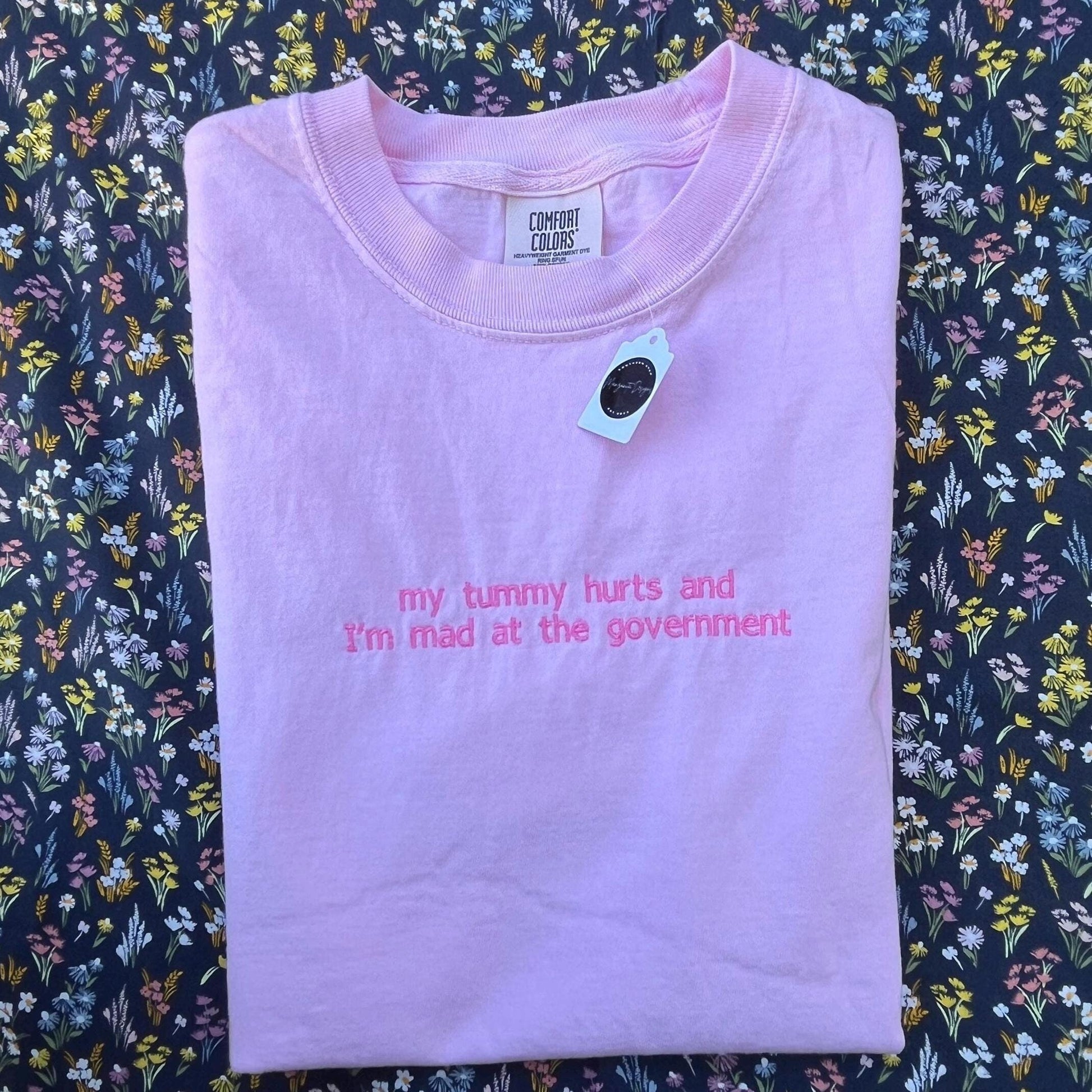 The image shows a folded light pink T-shirt with the text “my tummy hurts and I’m mad at the government” printed in small pink letters on the front. The shirt is placed on a floral-patterned fabric background with various small, colorful flowers