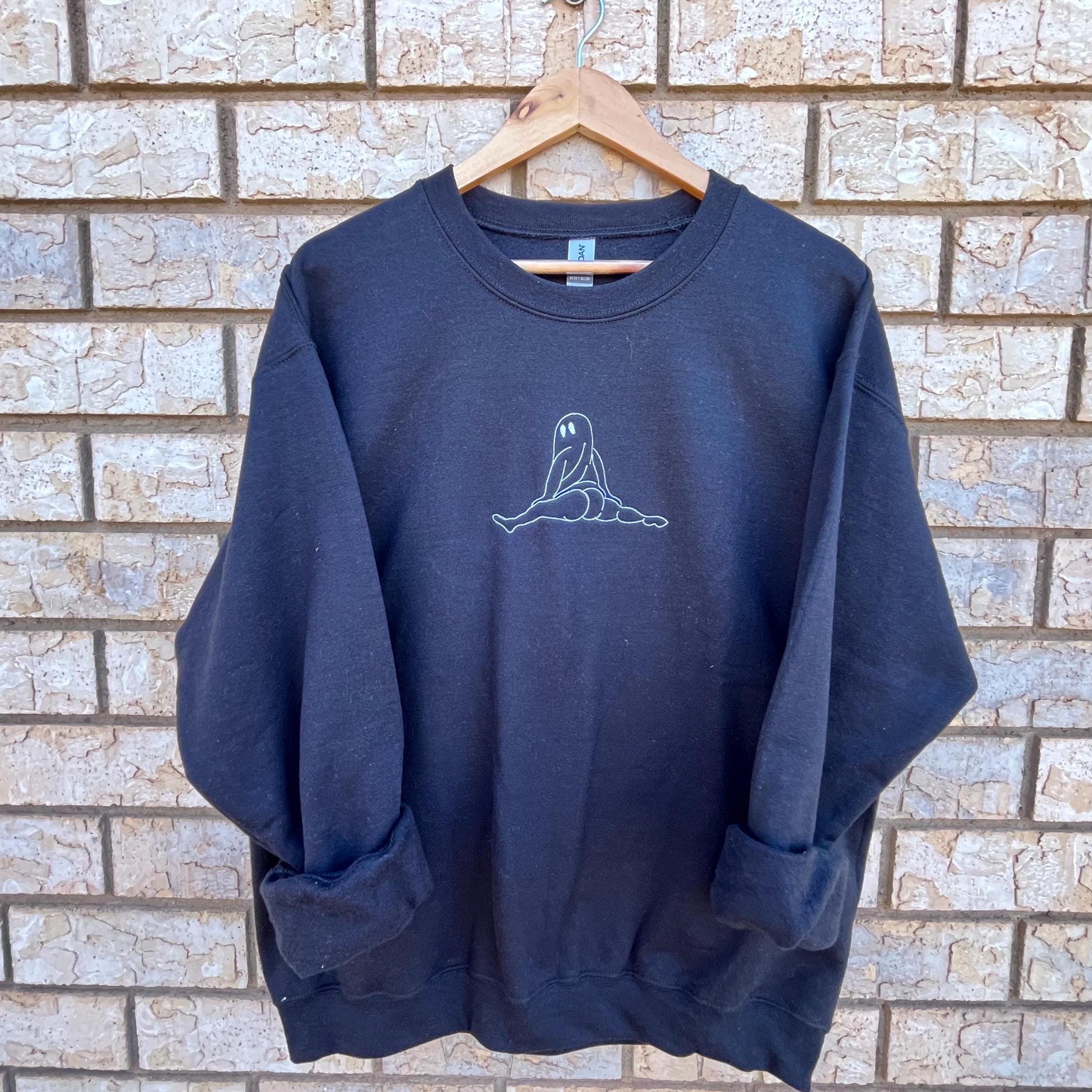 There's a black sweatshirt hanging on a wooden hanger against a brick wall. The sweatshirt features a simple line drawing of a seated figure. The figure is a ghost twerking. The drawing is located on the upper front part of the sweatshirt.