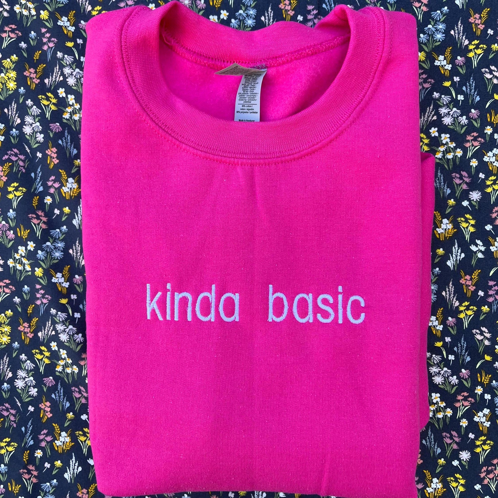 The image shows a folded bright pink sweatshirt with the words &quot;kinda basic&quot; embroidered in white on the front. The sweatshirt is placed on a floral-patterned fabric background with small, colorful flowers.