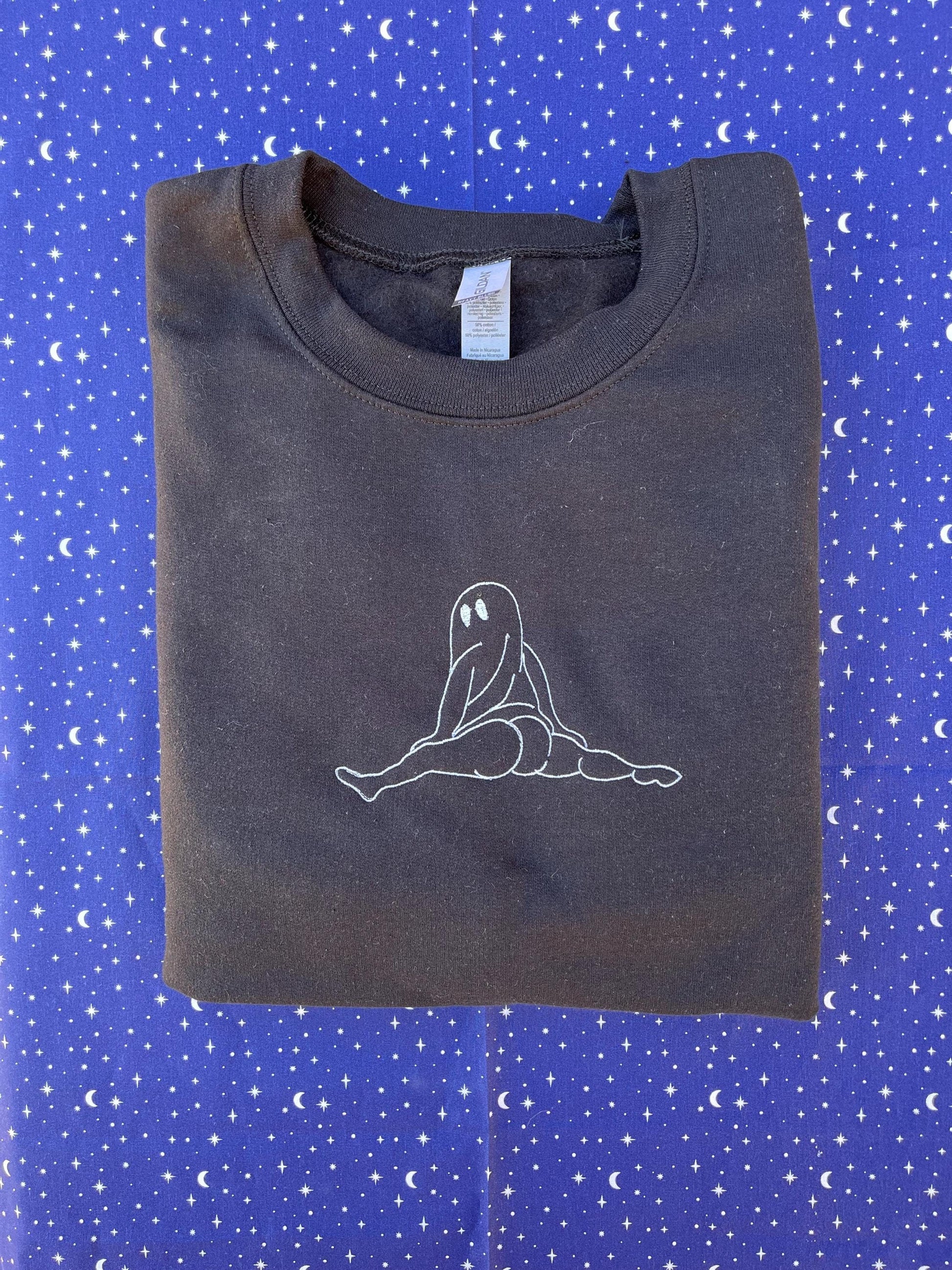 There's a black folded sweatshirt against a starry background. The sweatshirt features a simple line drawing of a seated figure. The figure is a ghost twerking. The drawing is located on the upper front part of the sweatshirt.